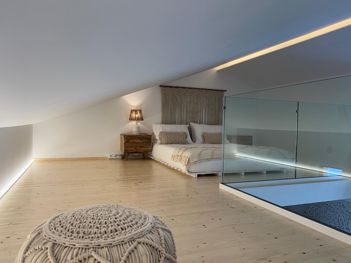 An open sleeping area is highlighted by a spacious bed with neutral linens. A decorative lamp rests on a wooden nightstand, and natural light filters through the sloped ceiling, enhancing the minimalist decor. A glass railing ensures safety while allowing views of the lower level.