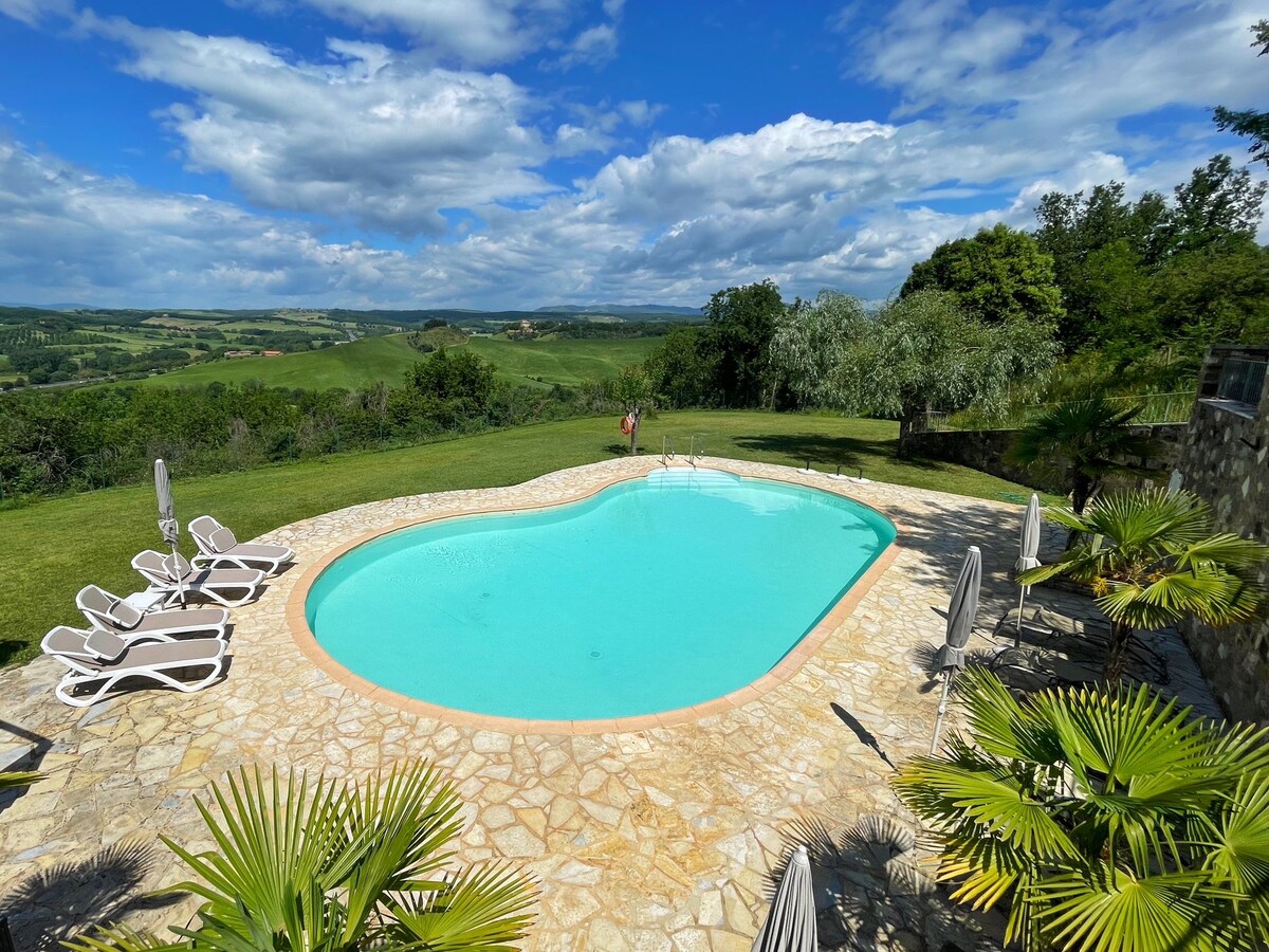 A serene pool area is set amidst lush greenery, featuring an irregularly shaped pool surrounded by sun loungers and umbrellas. The scenic countryside and distant hills create a calming backdrop beneath a partly cloudy sky.