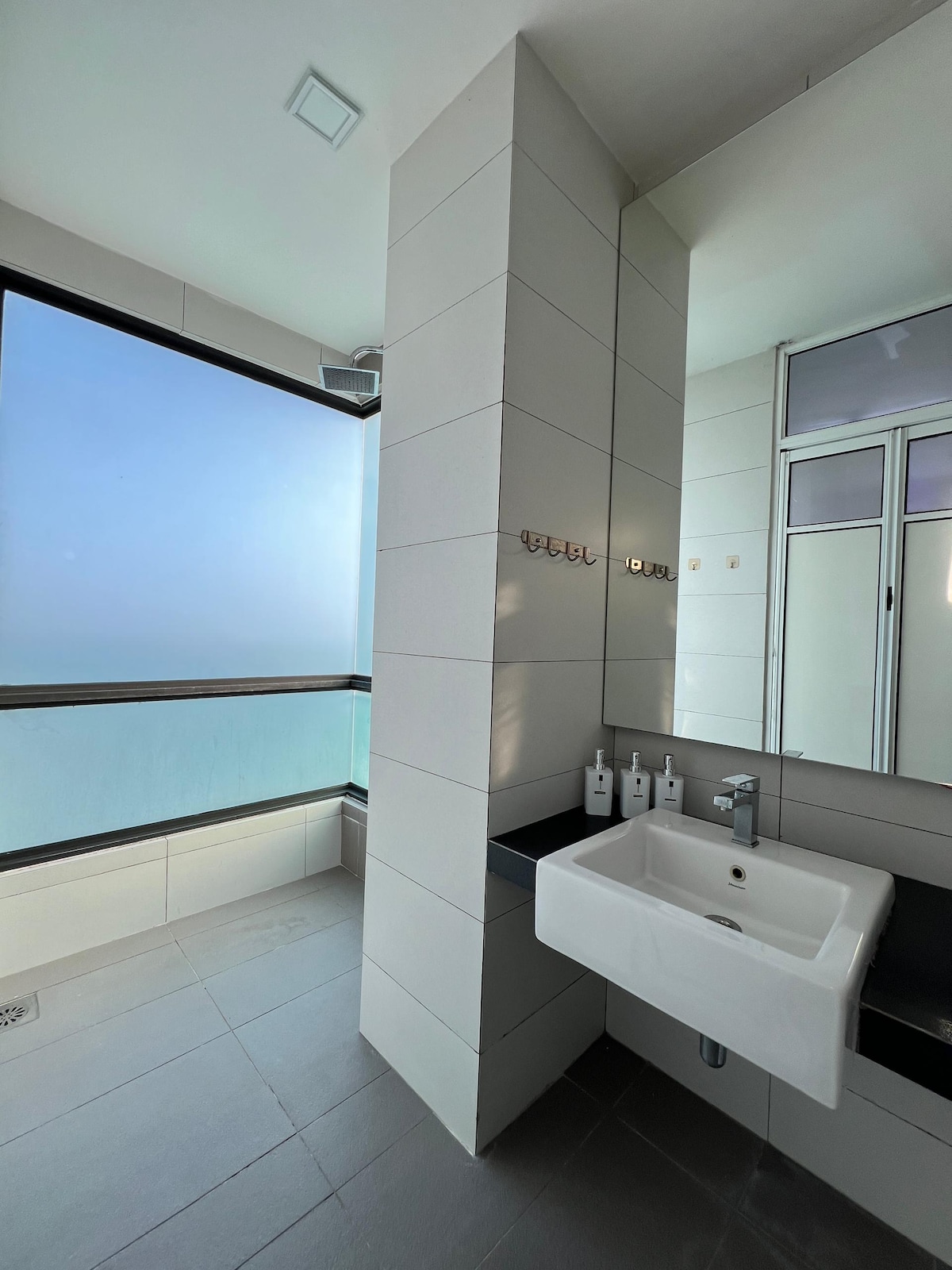 A modern bathroom is featured, with large windows allowing natural light to fill the space. A sleek sink and faucet are positioned on a dark countertop. Three matching bottles of toiletries are neatly placed beside the sink. Light-colored tiles create a clean and fresh atmosphere.
