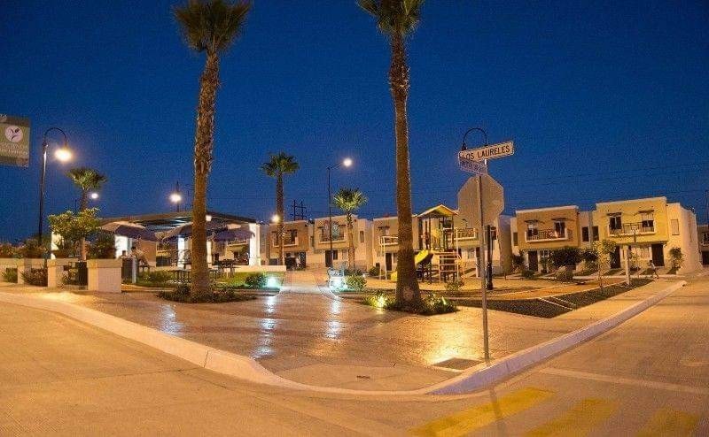 The outdoor area is illuminated by streetlights, highlighting well-maintained landscaping and palm trees. A playground is visible, accompanied by a cluster of modern residential buildings. The scene reflects a quiet atmosphere during nighttime with paved pathways winding through the space.
