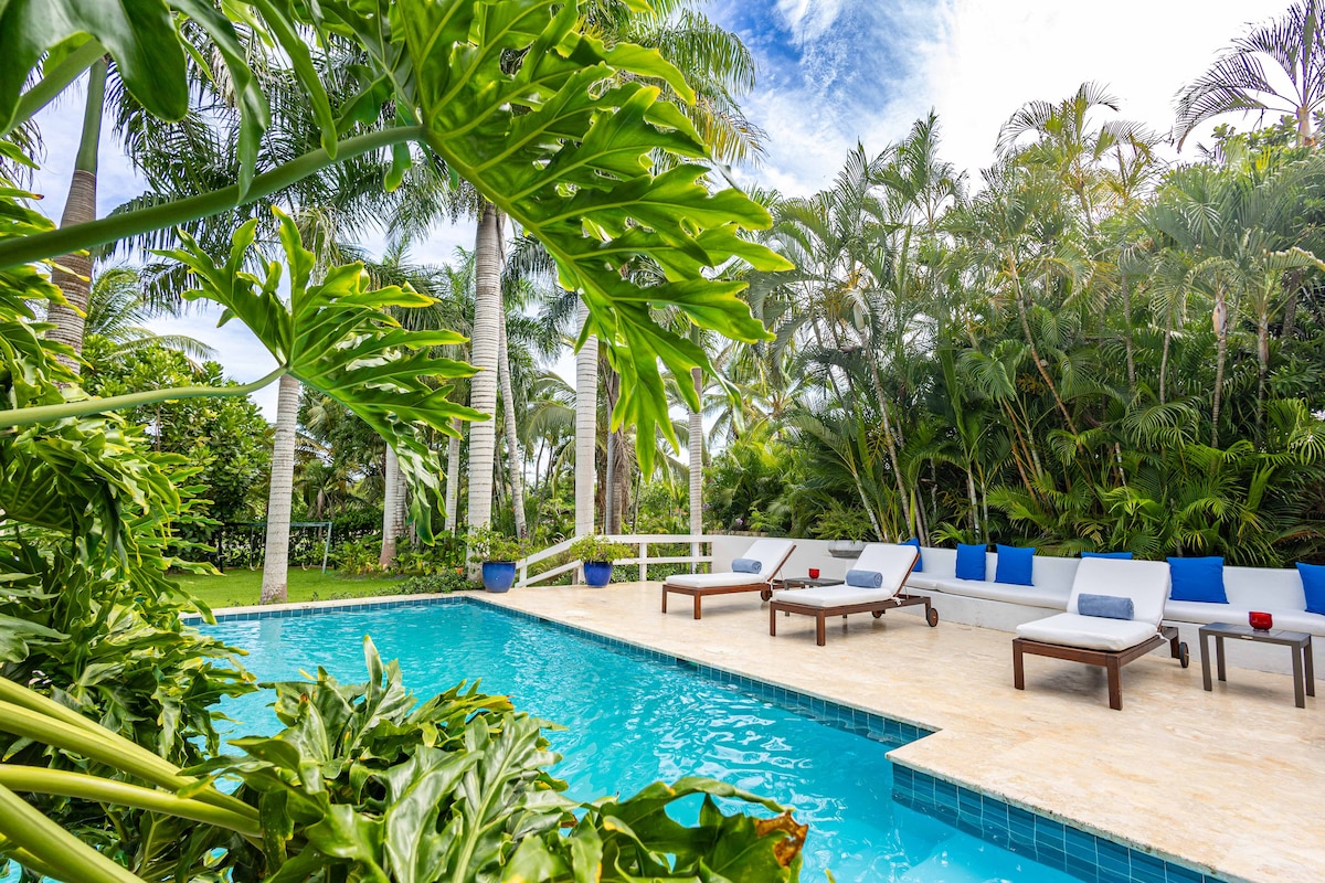 A inviting outdoor pool area is surrounded by lush tropical greenery, creating a serene atmosphere. Sun loungers are positioned nearby, providing a comfortable space for relaxation. The pool's clear water reflects the light, offering a refreshing escape. Palm trees gently sway in the background.