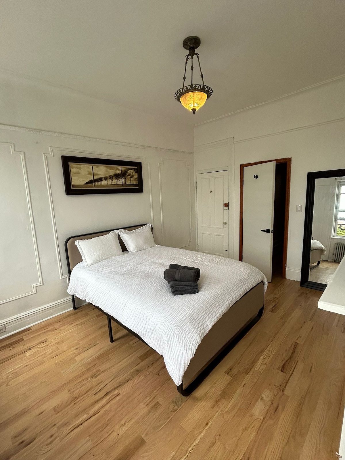 A bright bedroom features a spacious bed dressed in white linens, accompanied by two white pillows. A small towel is neatly arranged on the bed. Natural light enters through a nearby window, while a full-length mirror adds depth to the room's ambiance.