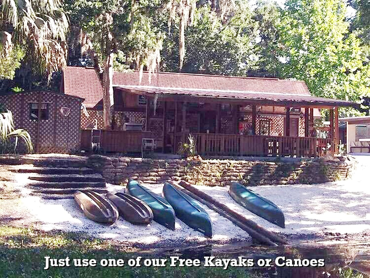 A charming cabin with cedar siding is situated beside a peaceful waterfront. Several canoes are arranged along a gravel path leading to the entrance, surrounded by lush greenery. The spacious deck offers a welcoming area for relaxation and outdoor activities.