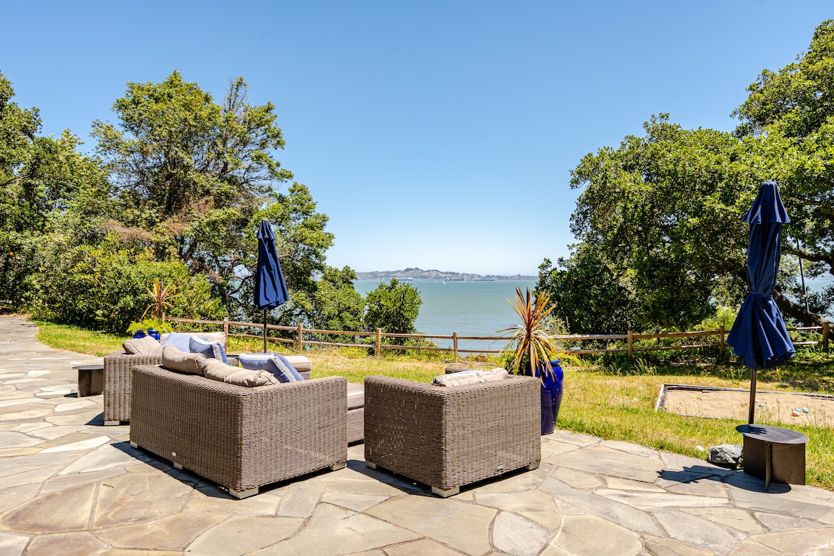 A seating area features comfortable outdoor couches, positioned on a stone patio. Umbrellas provide shade, and the view reveals serene water and distant hills, framed by lush greenery. The setting offers a relaxing spot for enjoying the natural surroundings.