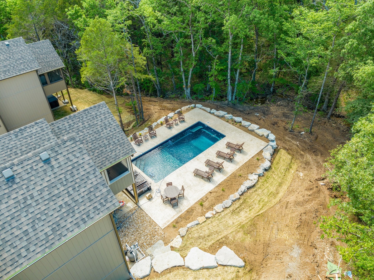 An aerial view captures a private saltwater pool surrounded by a stone patio. Lounge chairs are positioned for relaxation under the shade of nearby trees. The landscape features well-maintained grass and a mix of natural elements, enhancing the tranquil outdoor setting.