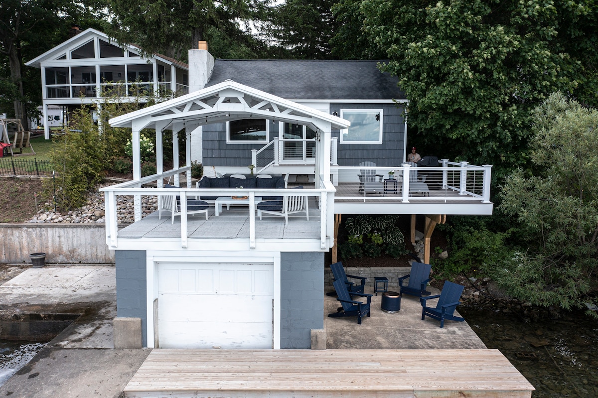 The image captures the exterior of a cozy cottage with a prominent boathouse deck. Comfortable outdoor seating is arranged around a fire pit on a spacious wooden deck, with the serene lakefront visible below. Lush greenery surrounds the structure, enhancing the tranquil lakeside setting.