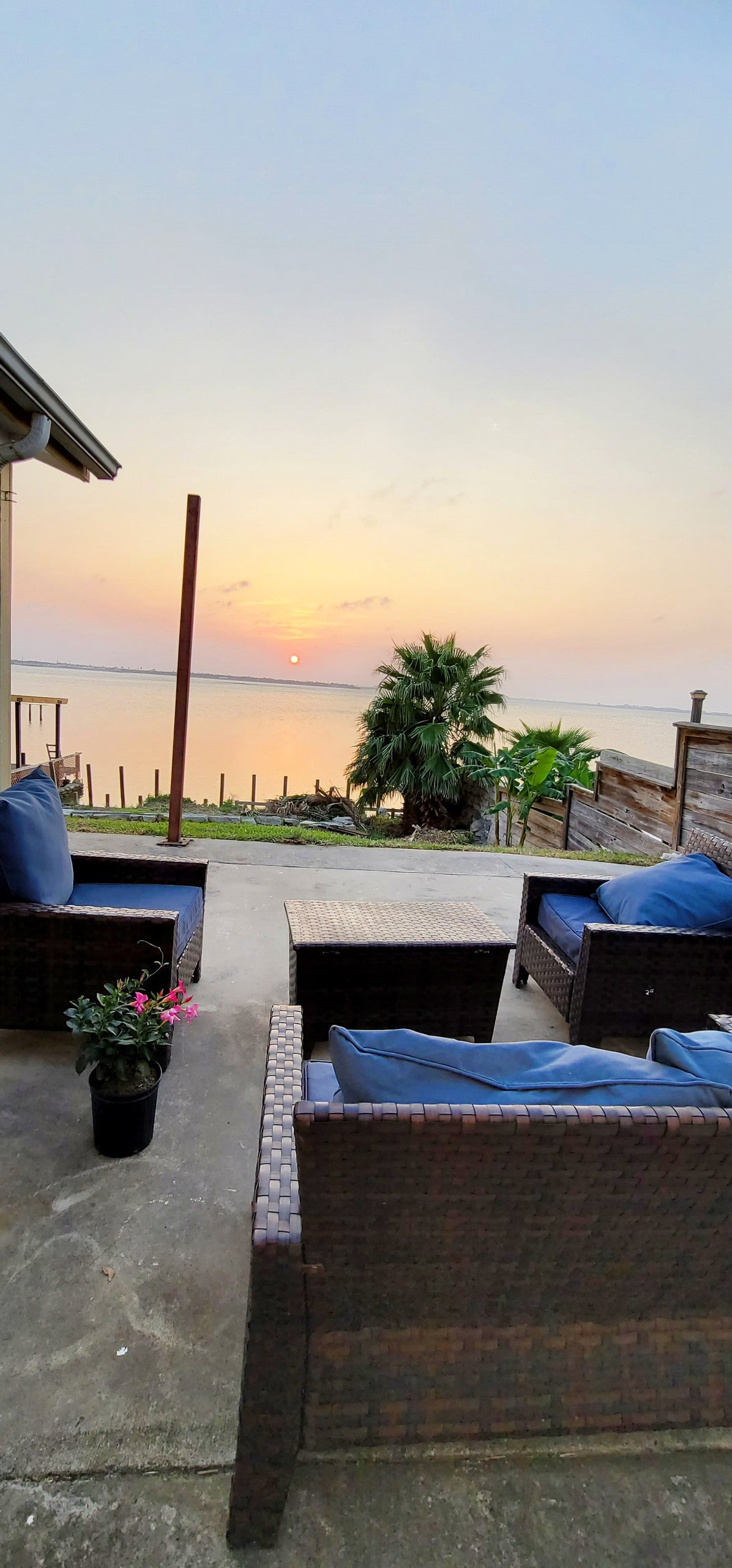 An outdoor seating area features woven seating with plush cushions, arranged around a small table. The scene is highlighted by a serene sunset reflecting on the water, with a palm plant and a flower pot adding a touch of greenery.