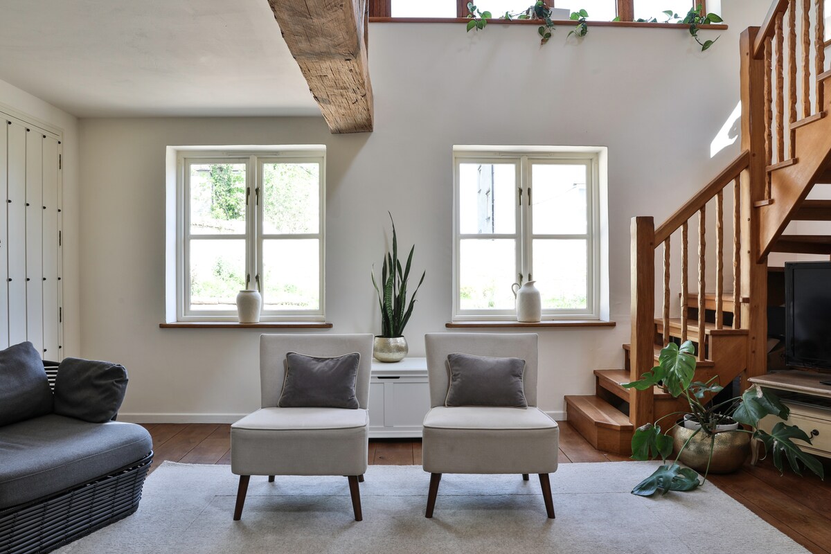 A bright living space features two stylish armchairs positioned on a light rug, with a staircase rising to the upper floor. Natural light enters through two large windows, where decorative pots and greenery are present. Original wooden beams add character to the room.