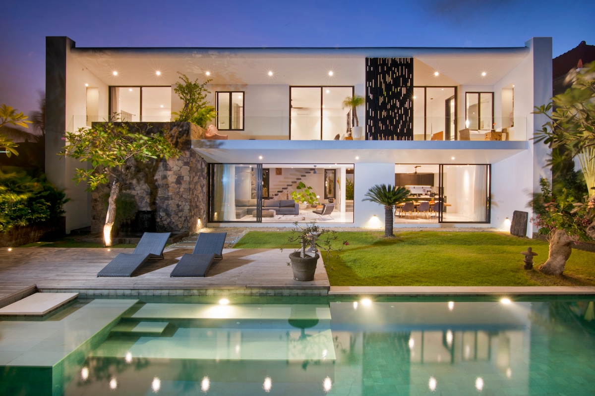 A modern villa is showcased with large glass windows that provide a seamless transition between indoor and outdoor spaces. The private pool reflects the surrounding greenery, while lounge chairs are positioned on the deck. Lush grass and tropical plants frame the exterior, enhancing the serene atmosphere.