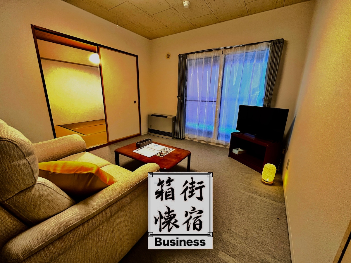 The living area features a comfortable seating arrangement with a light-colored sofa and a small wooden table. A flat-screen TV is positioned next to a window draped with gray curtains, allowing soft light to filter through. Traditional sliding doors lead to a tatami room.