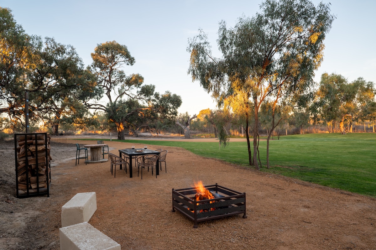 An outdoor area features a campfire/fire pit surrounded by a gravel surface. Nearby, a set of tables and chairs invites seating. Lush green grass stretches across the background, dotted with trees that enhance the natural setting.