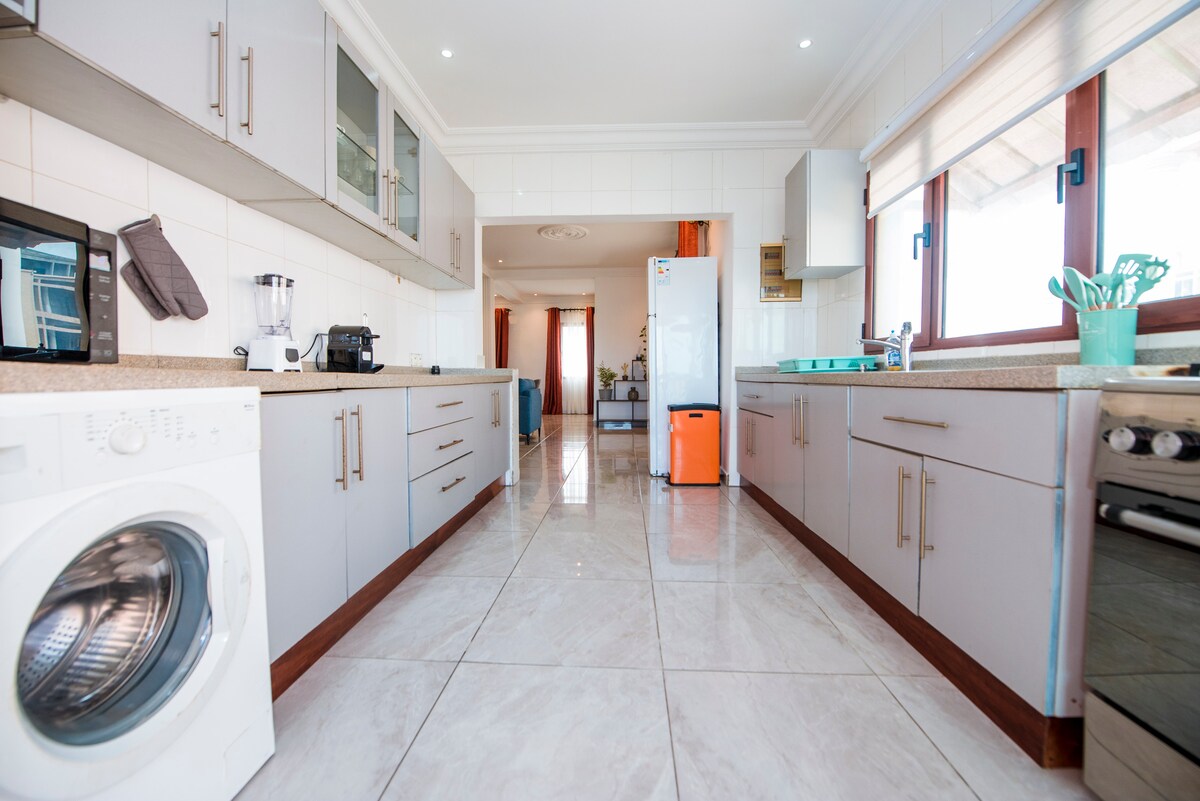 The kitchen features modern cabinetry and stainless steel appliances, including a refrigerator and a stove. A washing machine is positioned near the entrance. Bright, natural light filters in through the windows, illuminating the tiled floor and creating a functional cooking space.