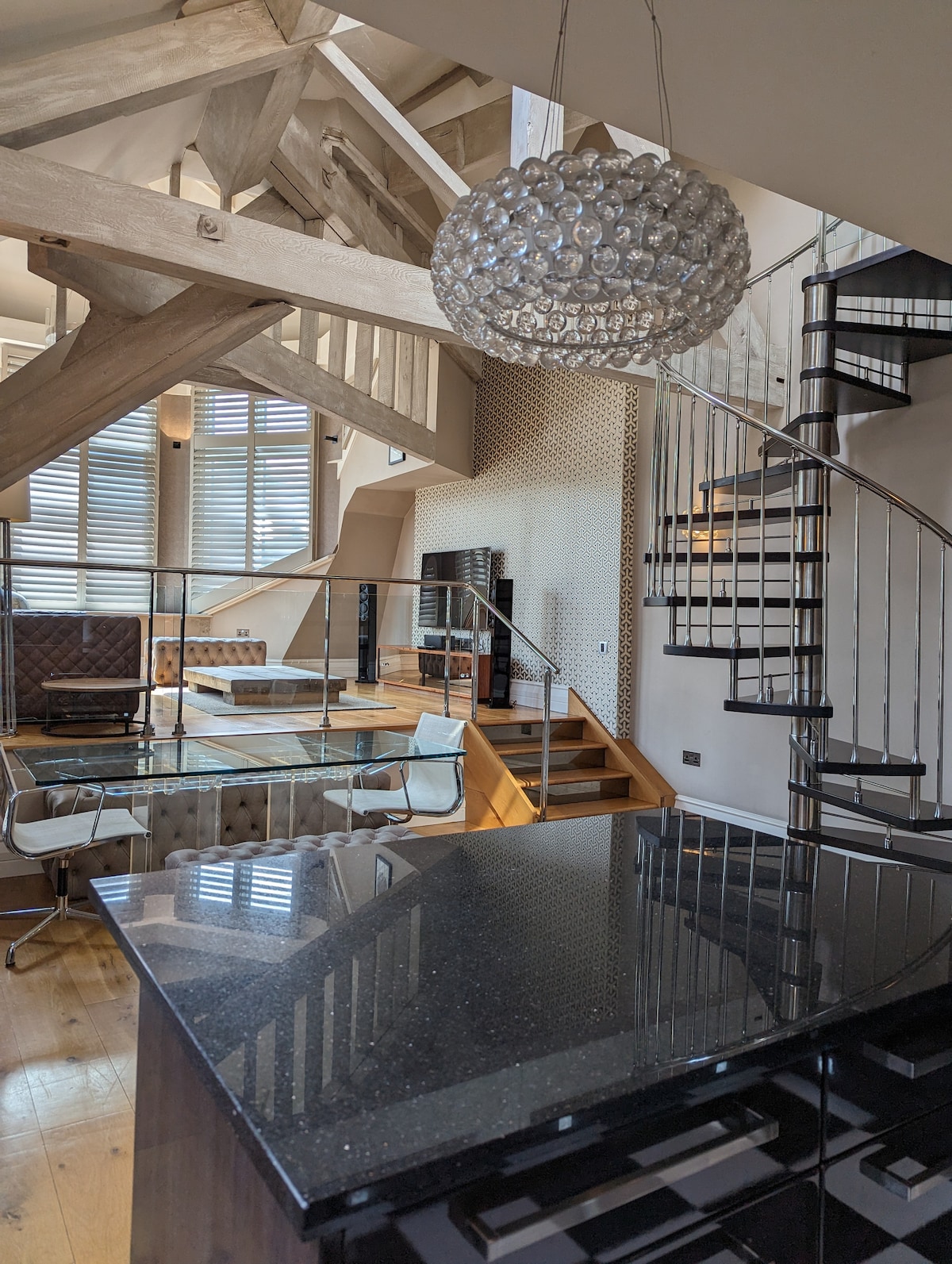 An open-concept living space features elegant lighting and a polished spiral staircase leading to the upper level. The room is adorned with large windows allowing natural light to illuminate the wooden flooring and modern furnishings, creating a welcoming atmosphere.