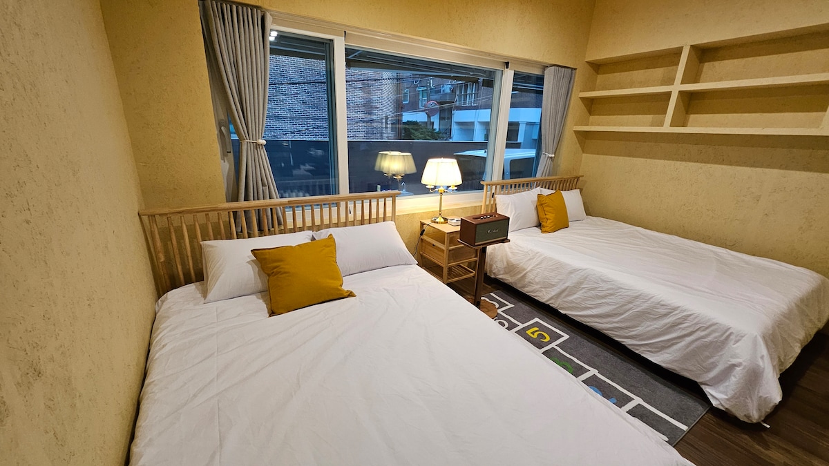 A bright sleeping area features two spacious beds positioned beside a large window. Natural light floods the room, accentuating the warm yellow walls. A decorative lamp and a small nightstand provide functional touches, while a patterned rug adds floor comfort.