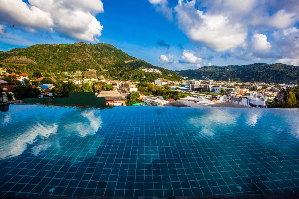 An infinity pool extends toward the horizon, reflecting the sky and surrounding mountains. Lush greenery and the town of Kata can be seen in the background, under a bright blue sky with scattered clouds, creating a serene view.