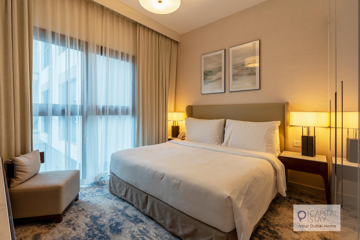 A well-appointed bedroom features a spacious bed with crisp white linens. Two bedside lamps provide soft lighting beside stylish wooden nightstands. Large windows with sheer curtains allow natural light to fill the room, while a cozy armchair sits in the corner on a textured area rug.
