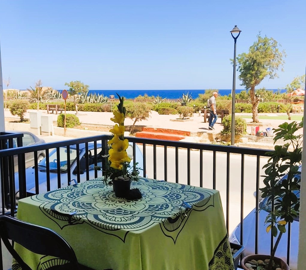 A sea view patio is showcased, featuring a vibrant table covered with a patterned cloth. Lush greenery surrounds the area, while the tranquil blue sea stretches out in the background. The scene captures the essence of relaxation and outdoor dining by the seaside.