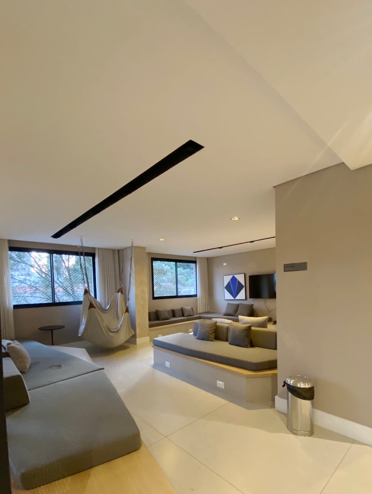 A spacious living area is highlighted by natural light filtering through large windows. A modern sofa arrangement is complemented by neutral-toned cushions. A hammock adds a relaxed touch, while a small side table and a colorful wall art piece contribute to the inviting atmosphere.