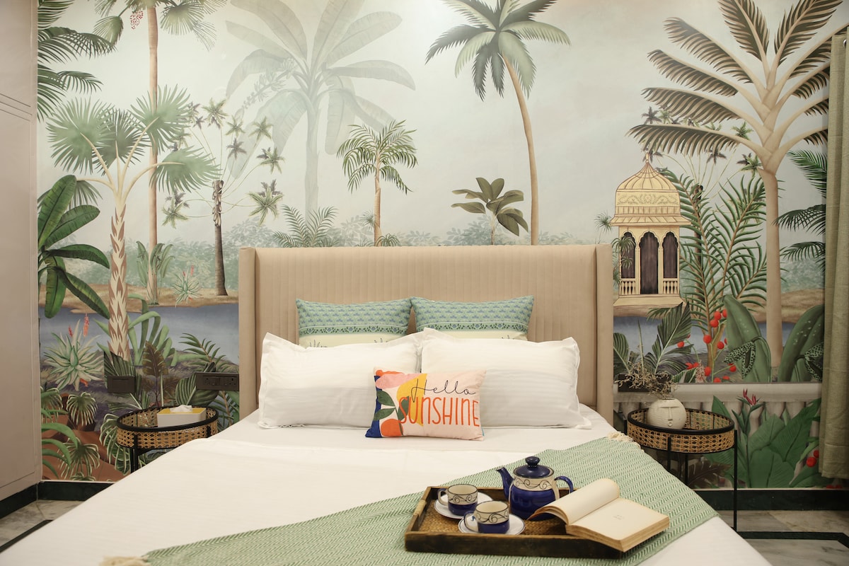 A tropical-themed bedroom features an expansive bed with a soft beige headboard. Wall murals of lush greenery enhance the space, complemented by stylish cushions. Decorative side tables hold cozy accents, while a vibrant pillow with the text 'Hello Sunshine' adds character to the overall aesthetic.