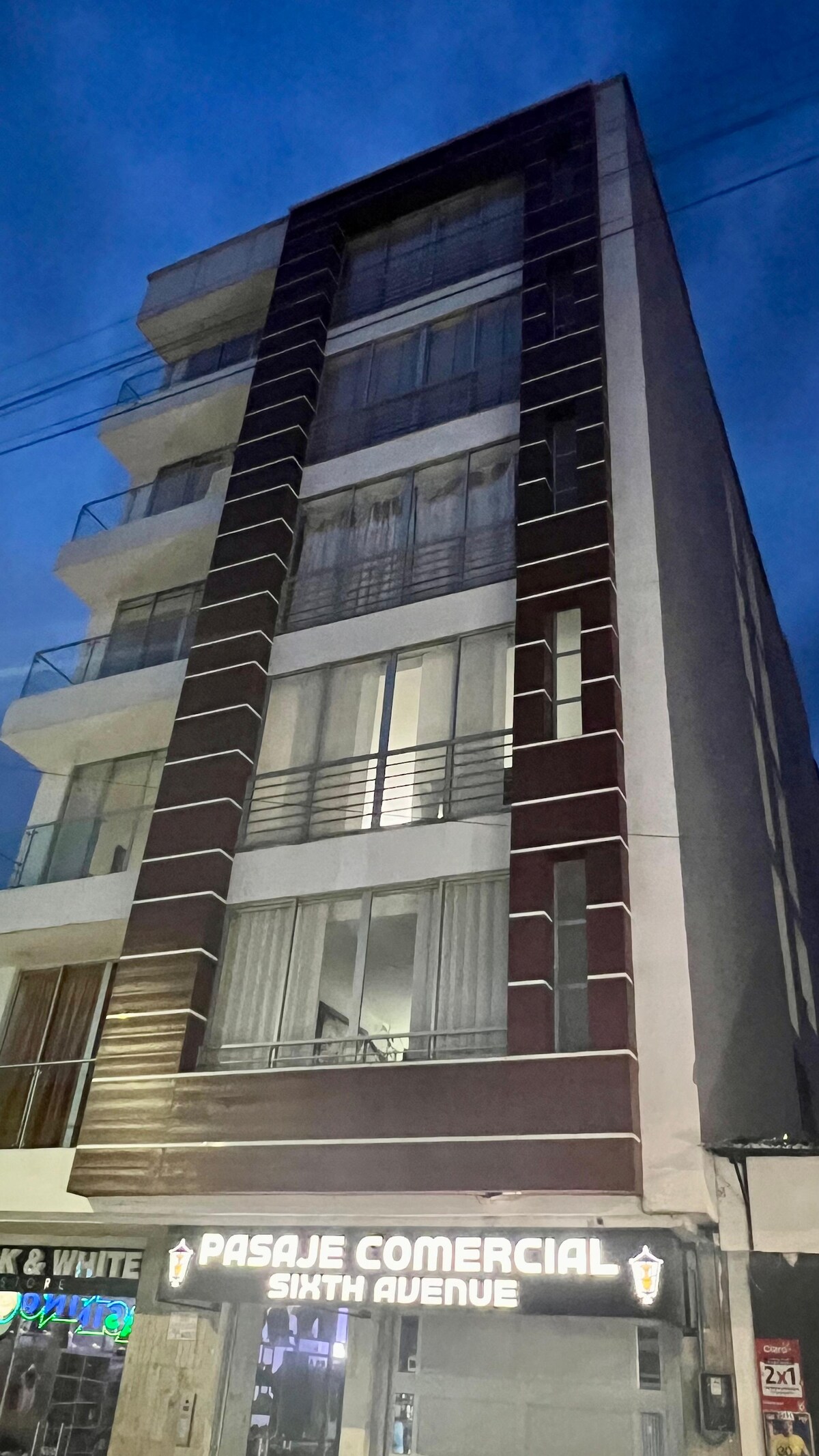 A modern five-story building is showcased in an evening setting. Bright lights from the commercial space illuminate the ground floor, while large windows above provide glimpses of the interior. The structure features a mix of light and dark facade elements, enhancing its contemporary appearance.