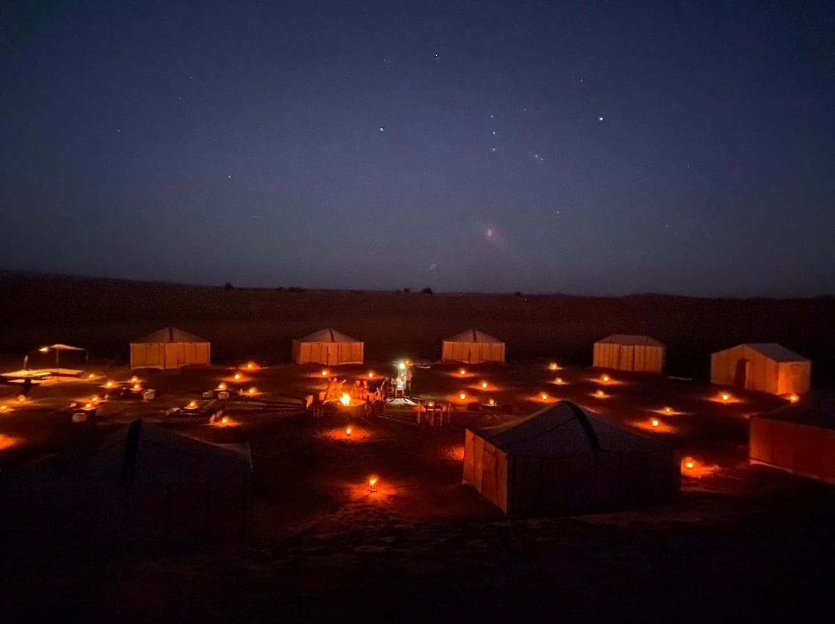 A desert camp is illuminated by soft orange lights, creating a serene nighttime atmosphere. Several tents are arranged in a circular formation, surrounded by dark sand and distant stars in the sky. A central gathering area is visible, featuring a fire pit inviting guests to relax.