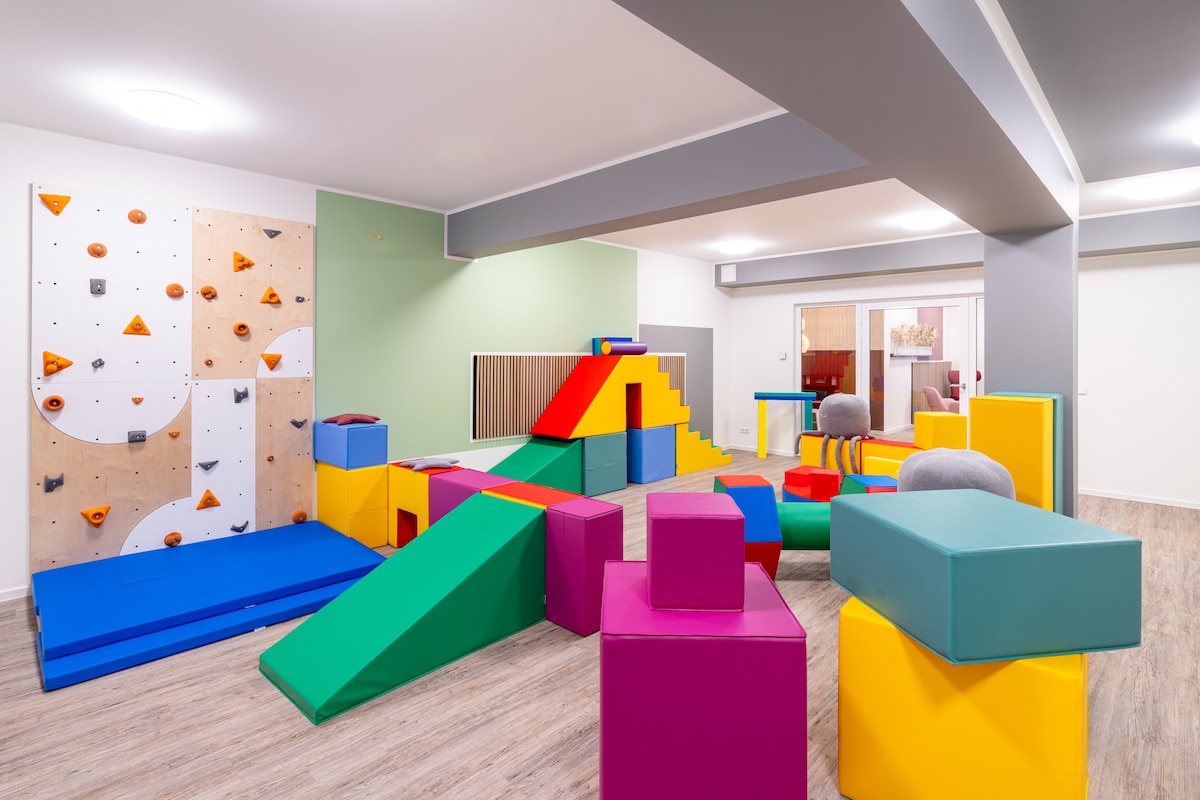 A colorful play area is equipped with various soft play equipment, including large blocks in vibrant colors and climbing structures. A small climbing wall is visible on one side, offering a safe space for children to explore and engage in physical activities.