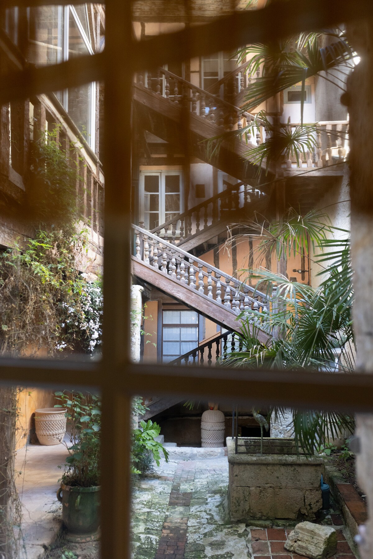 A view through a window frames an interior courtyard adorned with greenery. A wooden staircase with decorative railing is visible, leading to upper levels. Natural light filters softly, illuminating the textured walls and stone floor, creating a serene and inviting atmosphere.