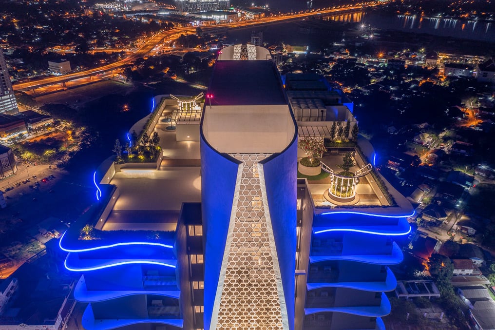 An aerial view captures the modern high-rise structure, illuminated by blue light. The rooftop showcases landscaped areas and stylish seating arrangements, while the cityscape sprawls below, revealing a network of roads and buildings, along with twinkling lights reflecting the vibrant atmosphere of Kota Bharu at night.