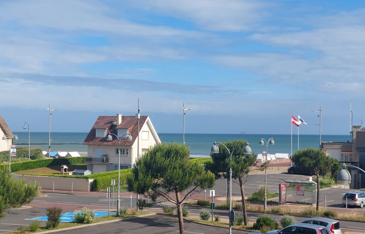 The image depicts a view of the sea from a distance, framed by a serene landscape. A house with a slanted roof is visible, along with green bushes and a few streetlights. The blue ocean meets a cloudy sky, suggesting a calm atmosphere.