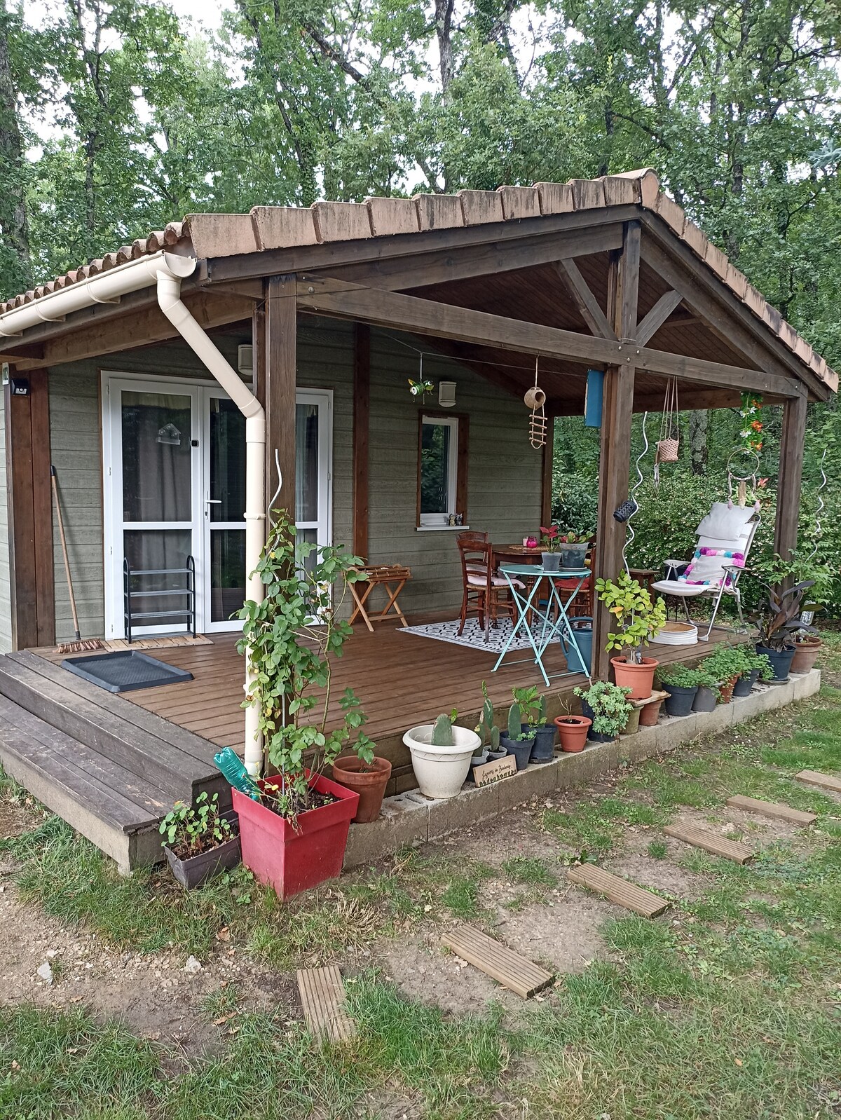 A charming chalet with a covered terrace is visible, surrounded by lush greenery. Colorful potted plants line the wooden deck, which features outdoor seating with a small table and chairs. The entrance is framed by clear glass doors leading into the interior.