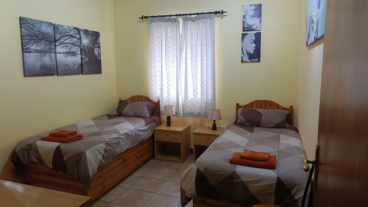 A twin bedroom featuring two wooden beds with a neutral color scheme. Each bed is adorned with patterned bedding and an orange towel. A small bedside table with lamps is positioned between the beds. Sunlight filters through a patterned curtain, illuminating the room's cheerful yellow walls.