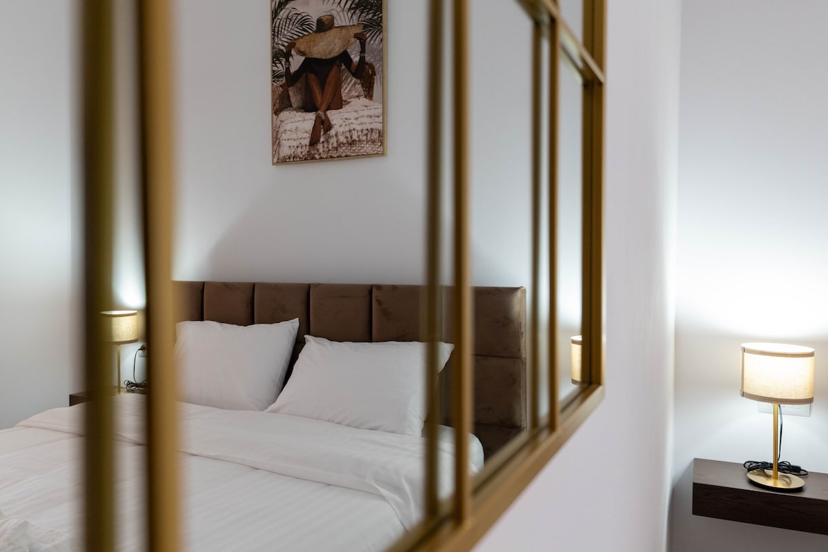 A well-appointed bedroom features a headboard with soft, neutral tones. Two bedside lamps cast gentle light, and a piece of wall art adds subtle character. The reflection in a decorative mirror creates depth, enhancing the warm atmosphere of the space.