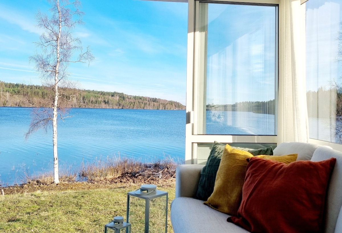 A sunlit sunroom is depicted with large glass windows overlooking a serene lake. A light sofa is adorned with colorful throw pillows. A small table is positioned beside the sofa, enhancing the tranquil lakeside setting.
