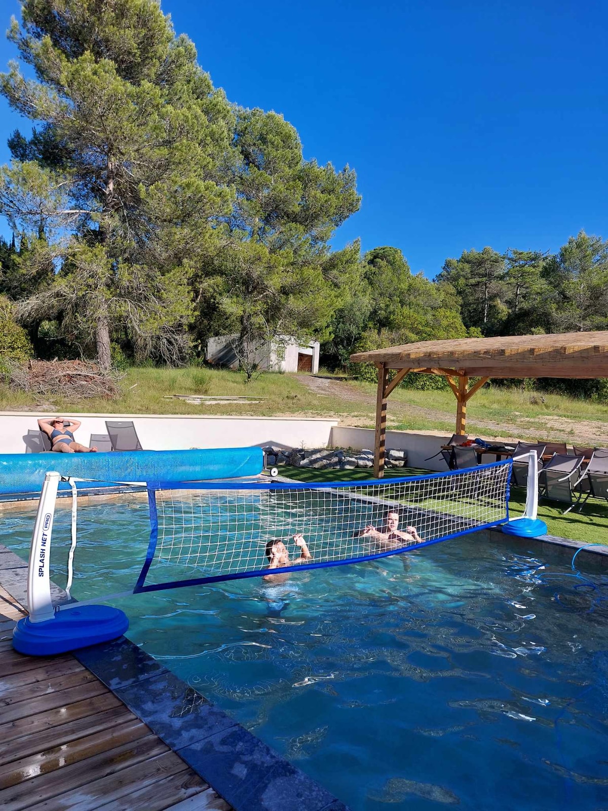 A spacious backyard pool is visible with swimmers enjoying a game of water volleyball. Sun loungers are positioned nearby under a wooden pavilion, surrounded by lush greenery. The clear blue sky provides a bright backdrop, and the pool is framed by wooden decking.