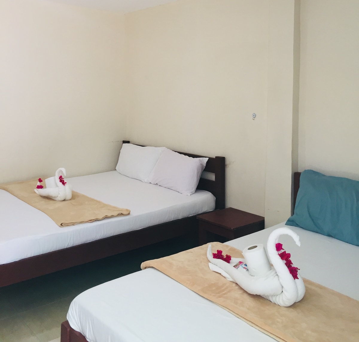 The room features three spacious beds arranged neatly. Elegant towel swans adorned with vibrant flowers are placed on the beds. A light neutral wall complements the simple furniture, including a small side table. Natural light streams through an unobtrusive window.