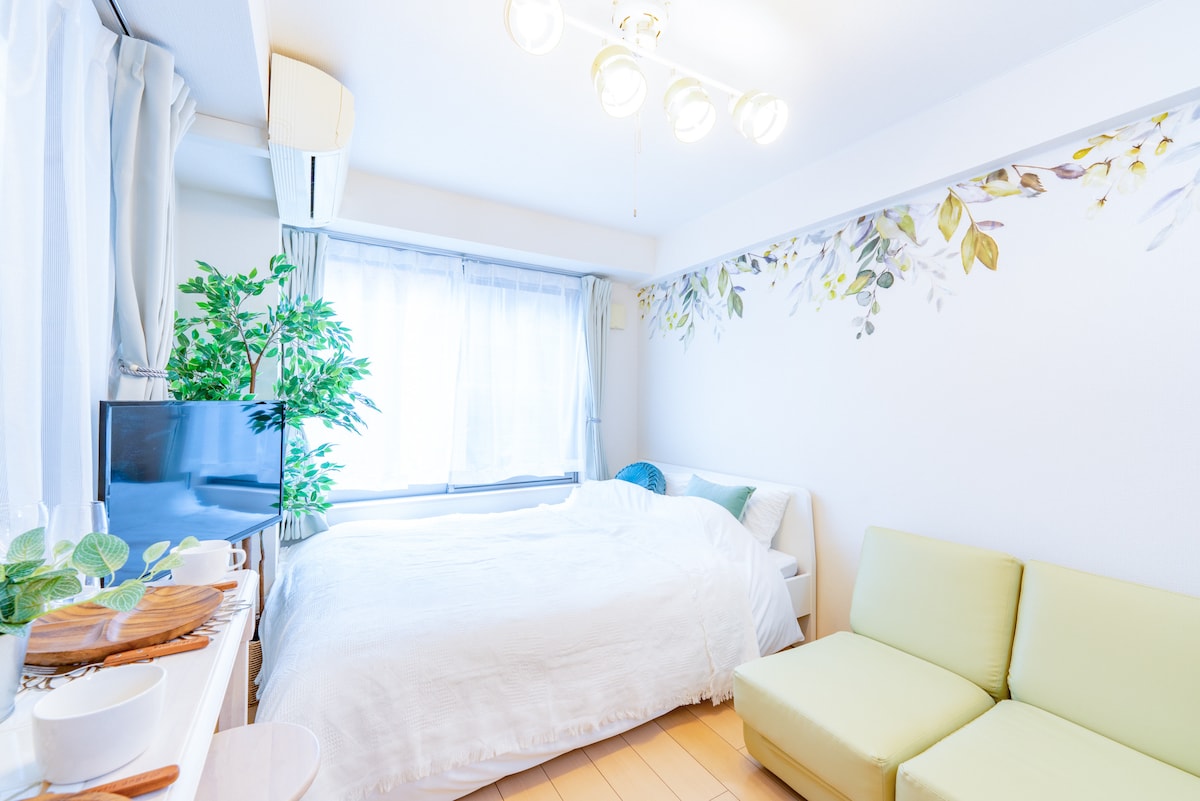 A simple and bright room is illuminated by natural light from a large window. A spacious bed is draped with a white coverlet, complemented by decorative pillows. A light green sofa adds a pop of color, while a television sits on a nearby shelf, surrounded by decorative plants.