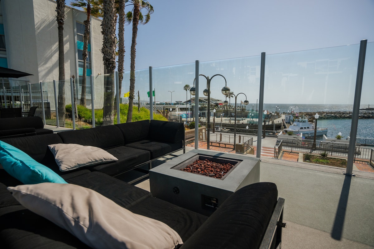 An outdoor seating area is shown featuring a black sectional sofa adorned with soft pillows in light tones. A fire pit with decorative stones is centered on a deck overlooking the ocean and marina, framed by palm trees and clear blue skies.