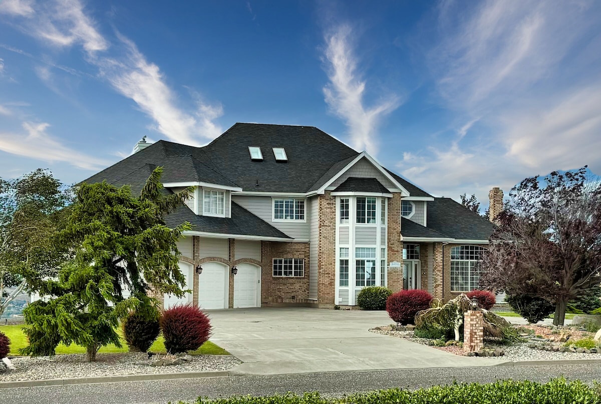 The serene exterior of a spacious hilltop house is presented, showcasing a two-story brick façade with multiple large windows. A well-maintained driveway leads to a double garage, surrounded by lush green foliage and colorful bushes. A scenic sky enhances the inviting atmosphere.