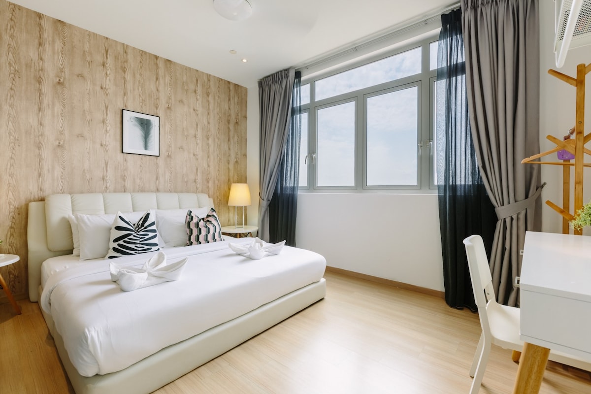 A well-lit bedroom features a spacious bed with decorative pillows and two neatly arranged towels. Large windows allow natural light to brighten the room, and sheer curtains add texture. A small desk and chair sit adjacent to the bed, enhancing the functional layout.