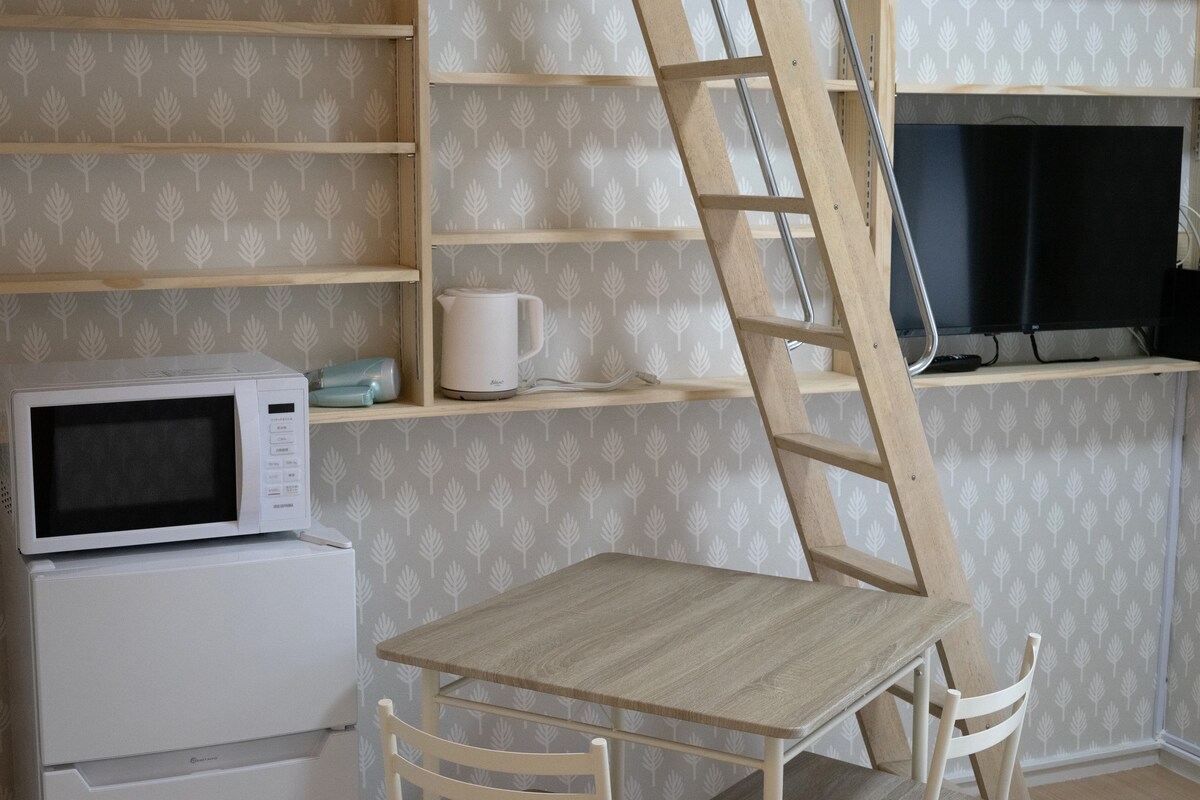 A minimalist living space includes a light wood dining table with two chairs and a television mounted on the wall. A compact microwave and a kettle are positioned on a shelf. A wooden ladder leads to the loft above, enhancing accessibility and creativity in use.
