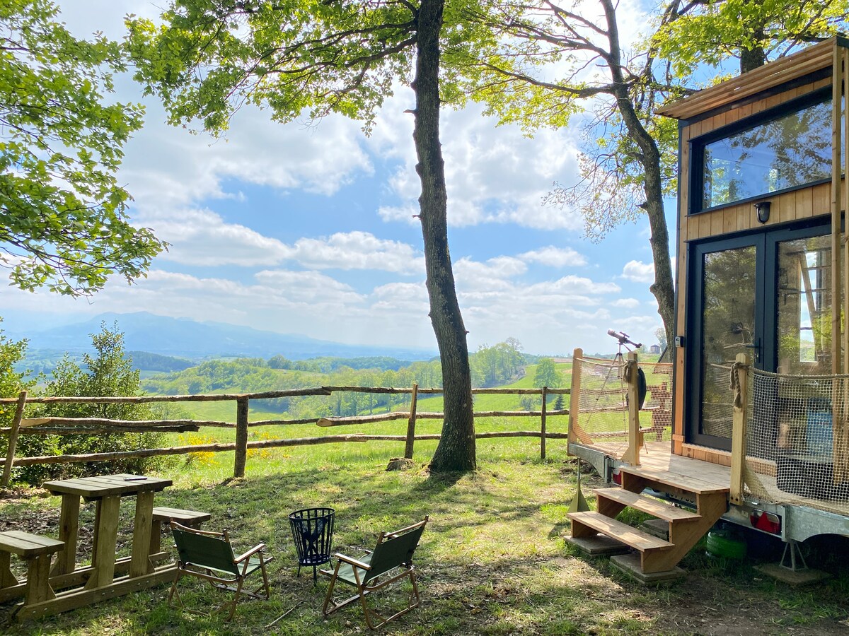An outdoor area features a wooden table and chairs near the entrance of the refuge. A fire pit is positioned on the grass, surrounded by trees. The scene opens to a panoramic view of rolling hills and a bright blue sky with scattered clouds.