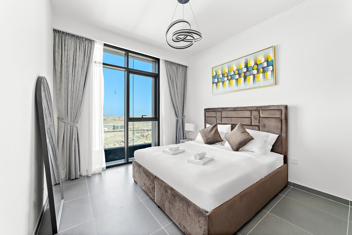 A well-appointed bedroom features a spacious bed with coordinated pillows, flanked by bedside lamps. Large windows provide abundant natural light and showcase the distant cityscape. A decorative mirror and modern art add visual interest to the room's neutral color scheme.