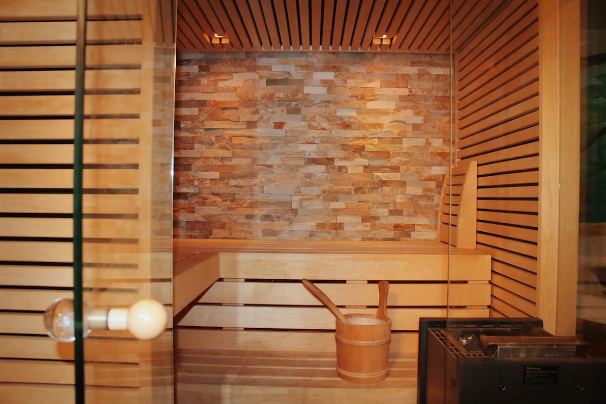 A serene indoor sauna is constructed with light wood and accented by stone walls. A wooden sauna bucket is placed on the floor near a heating unit, and soft lighting enhances the calming atmosphere.