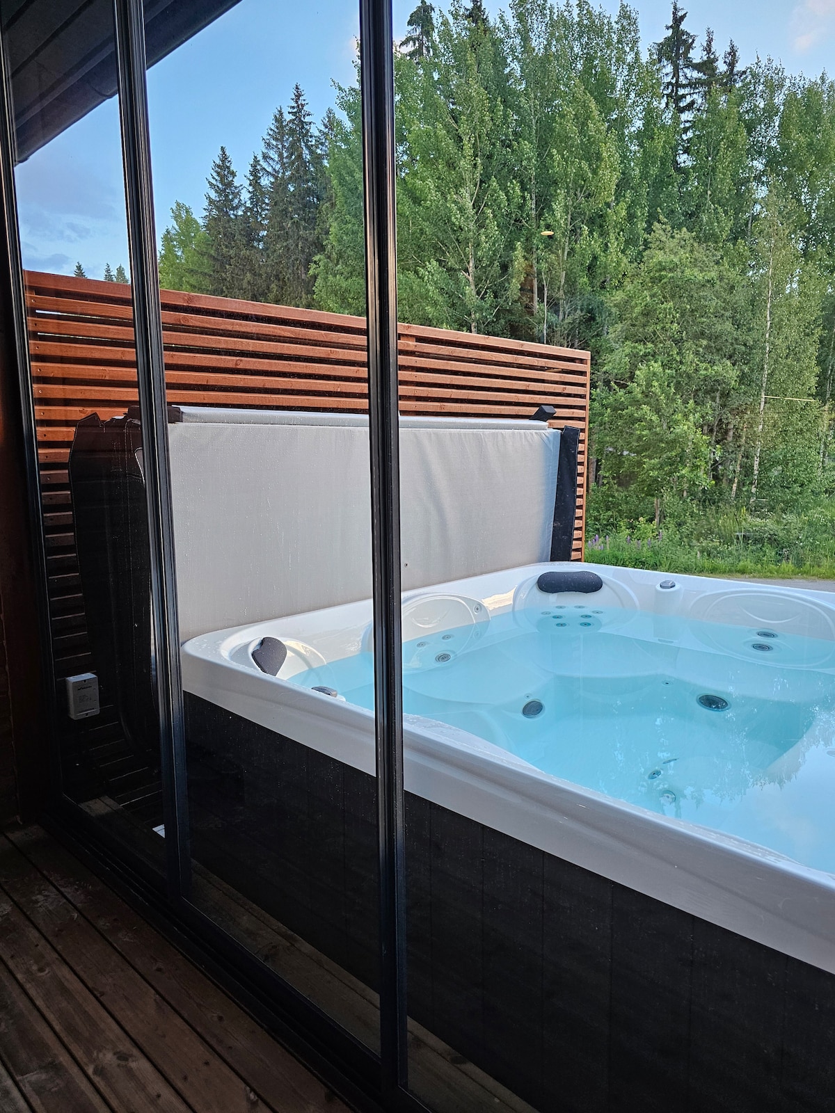 An outdoor hot tub is positioned on a wooden deck, framed by glass panels. The water appears inviting and is surrounded by lush green trees. A privacy screen is partially lowered, ensuring a serene atmosphere.