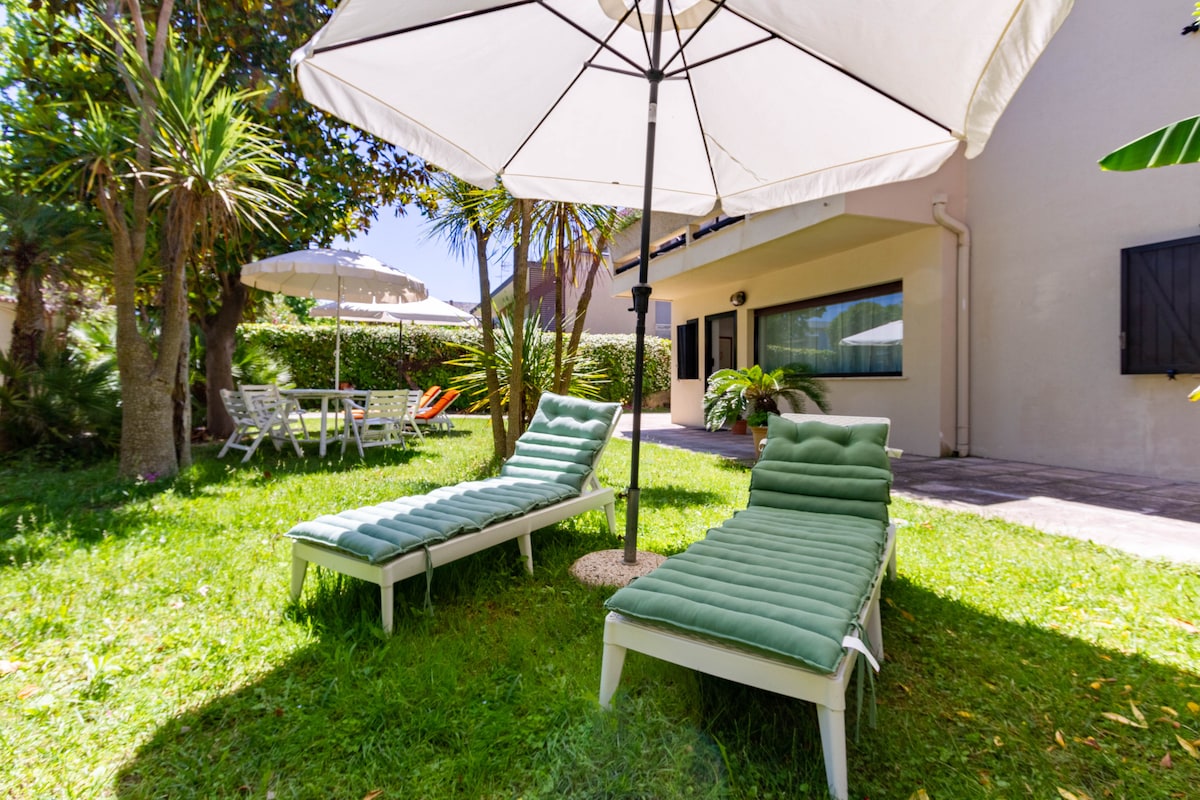 Two sun loungers with green cushions are positioned on a grassy area, shaded by a large umbrella. In the background, a dining table and chairs are visible, surrounded by lush greenery, creating a relaxing outdoor atmosphere.