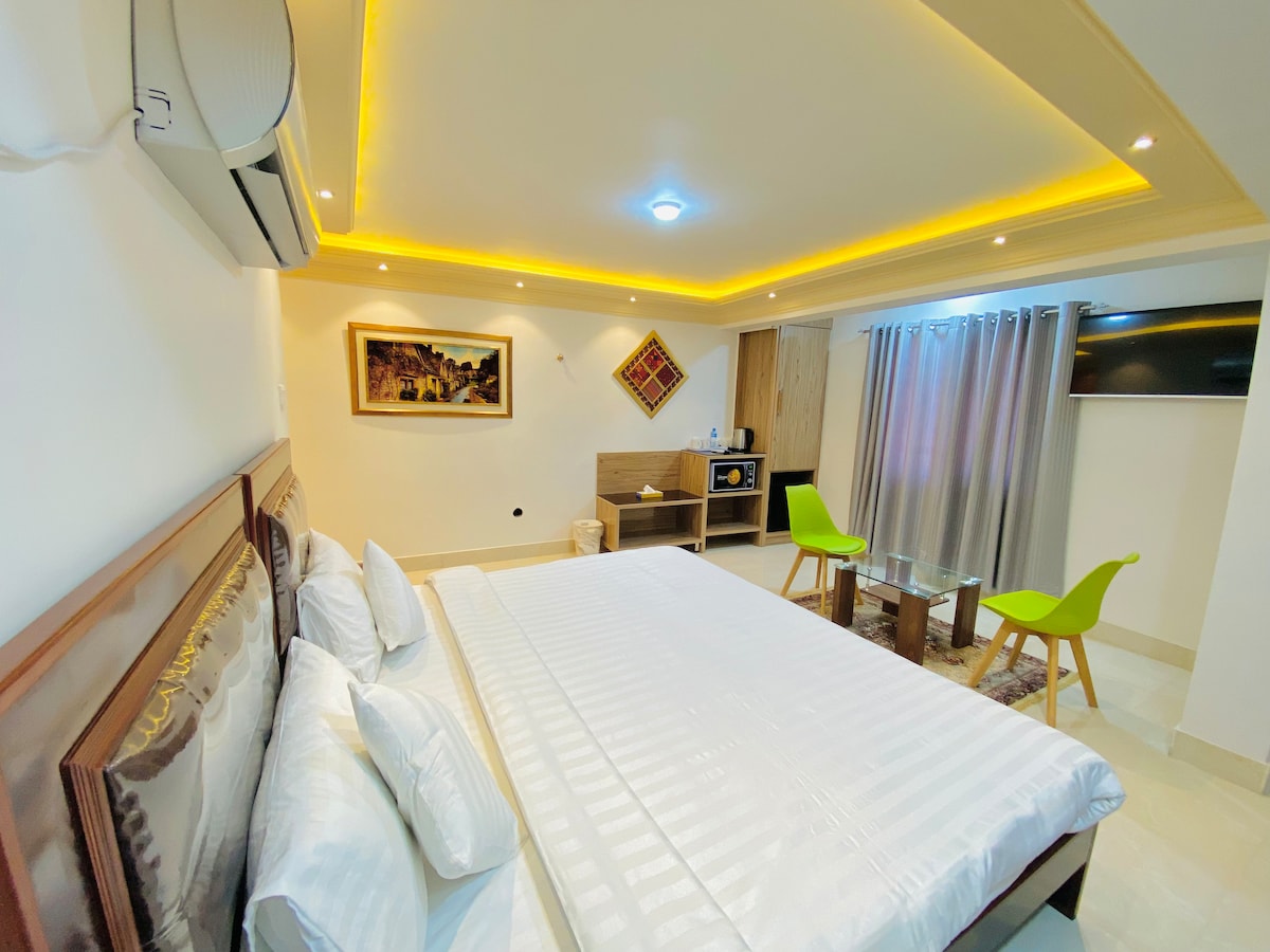 The well-lit room showcases a spacious layout featuring a neatly made bed with white linens. A flat-screen TV is mounted on the wall, while a small table and two chairs provide seating. A microwave and fridge are available, with curtains partially drawn to allow natural light.