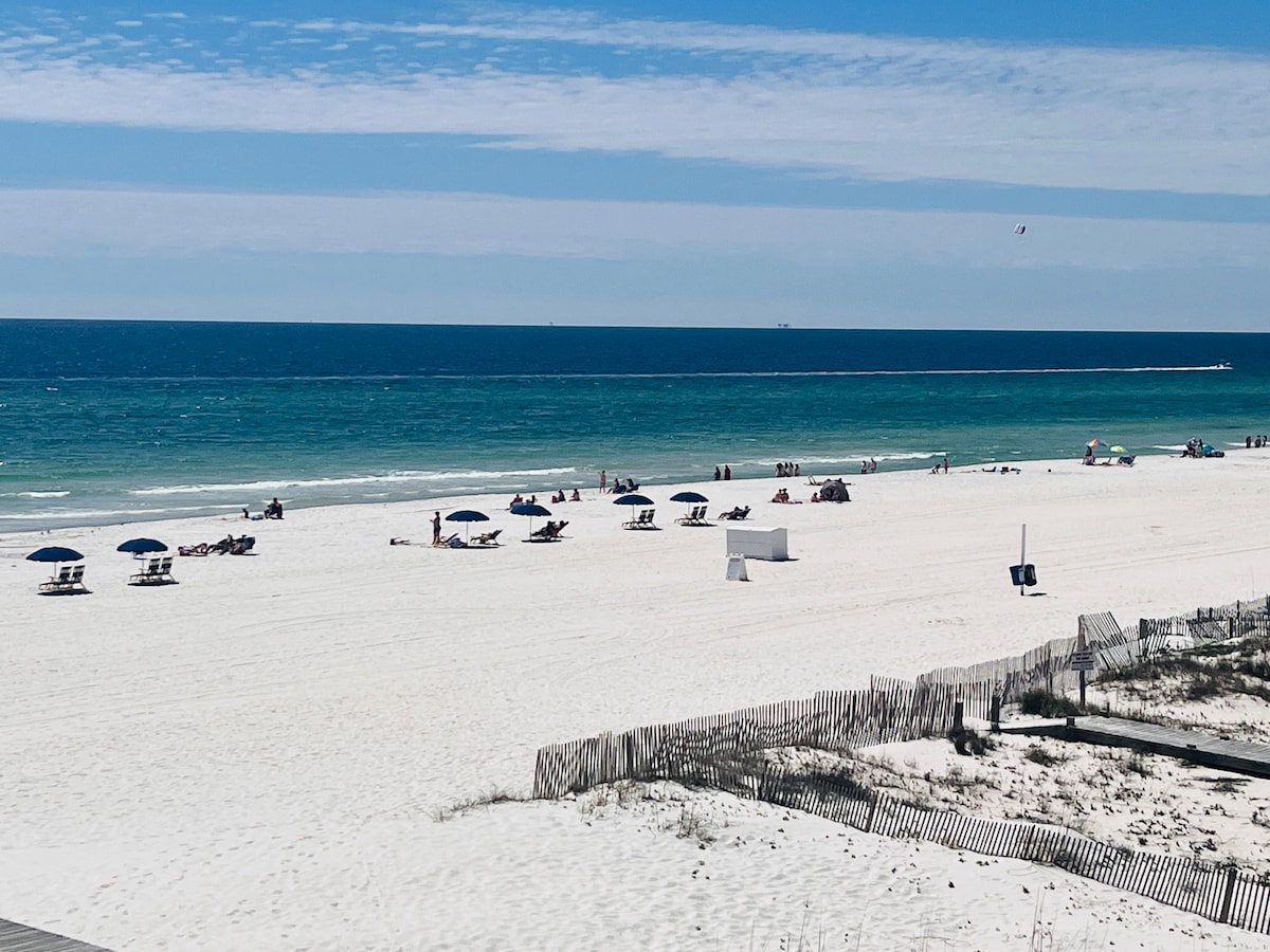 A serene beach scene is displayed, featuring soft white sand and clear turquoise waters under a bright blue sky. Several beach umbrellas and lounge chairs are positioned along the shore, while distant figures enjoy the sun. A natural dune fence borders the beach area.