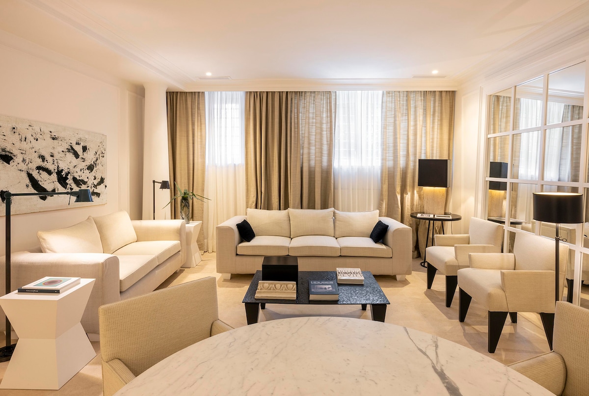 A spacious living area features light-colored sofas arranged around a low coffee table. Soft, natural light filters through large windows draped with elegant curtains. Two armchairs complement the seating, while a sleek floor lamp and stylish side tables enhance the inviting atmosphere.