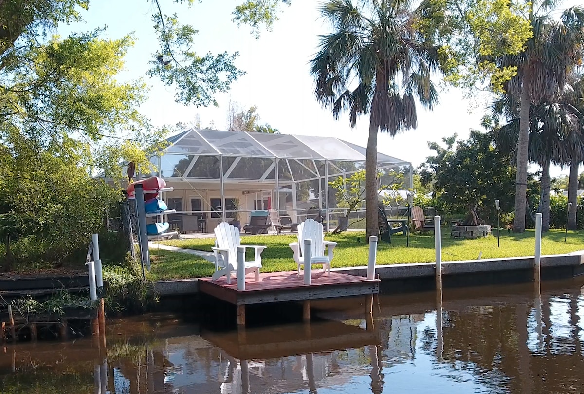 A private dock featuring two white Adirondack chairs overlooks the calm waters of the creek. Lush greenery surrounds the area, with a screened-in pool area visible in the background, offering a serene setting for relaxation and outdoor enjoyment.