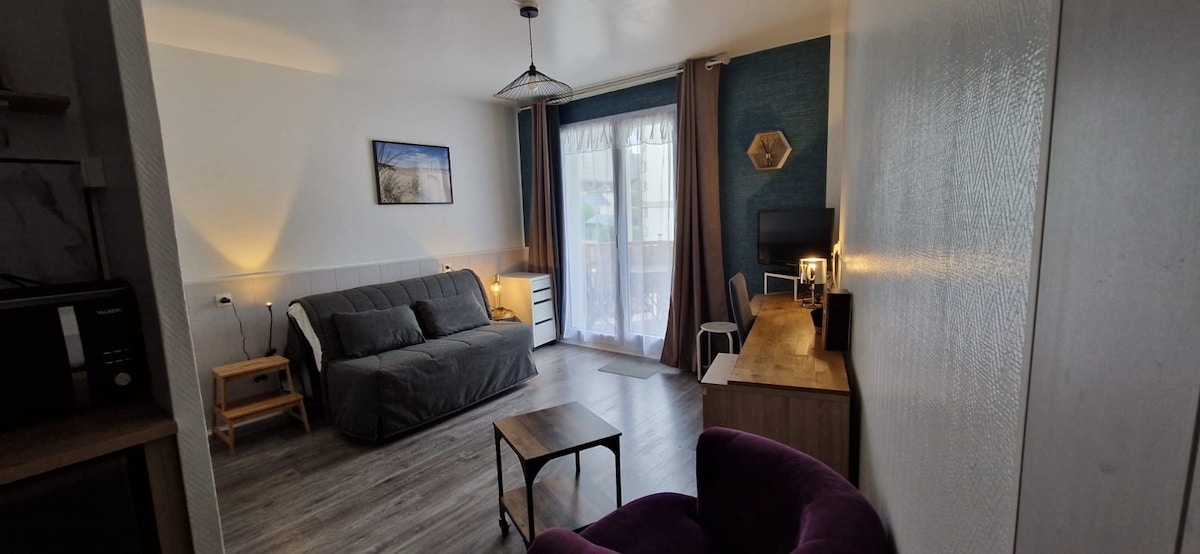 A modern studio living area features a grey sofa, a wooden coffee table, and a workspace with a chair. Natural light floods in from the balcony door, which is partially covered by sheer curtains. A flat-screen TV and decorative wall art enhance the ambiance.